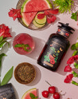 A jar of Watermelon Mint Herbal Iced Tea is surrounded by a glass of iced tea, sliced watermelon, cherries, lime slices, red flowers, and a bowl of loose tea leaves with a hint of organic peppermint. The setup is laid out on a white surface.