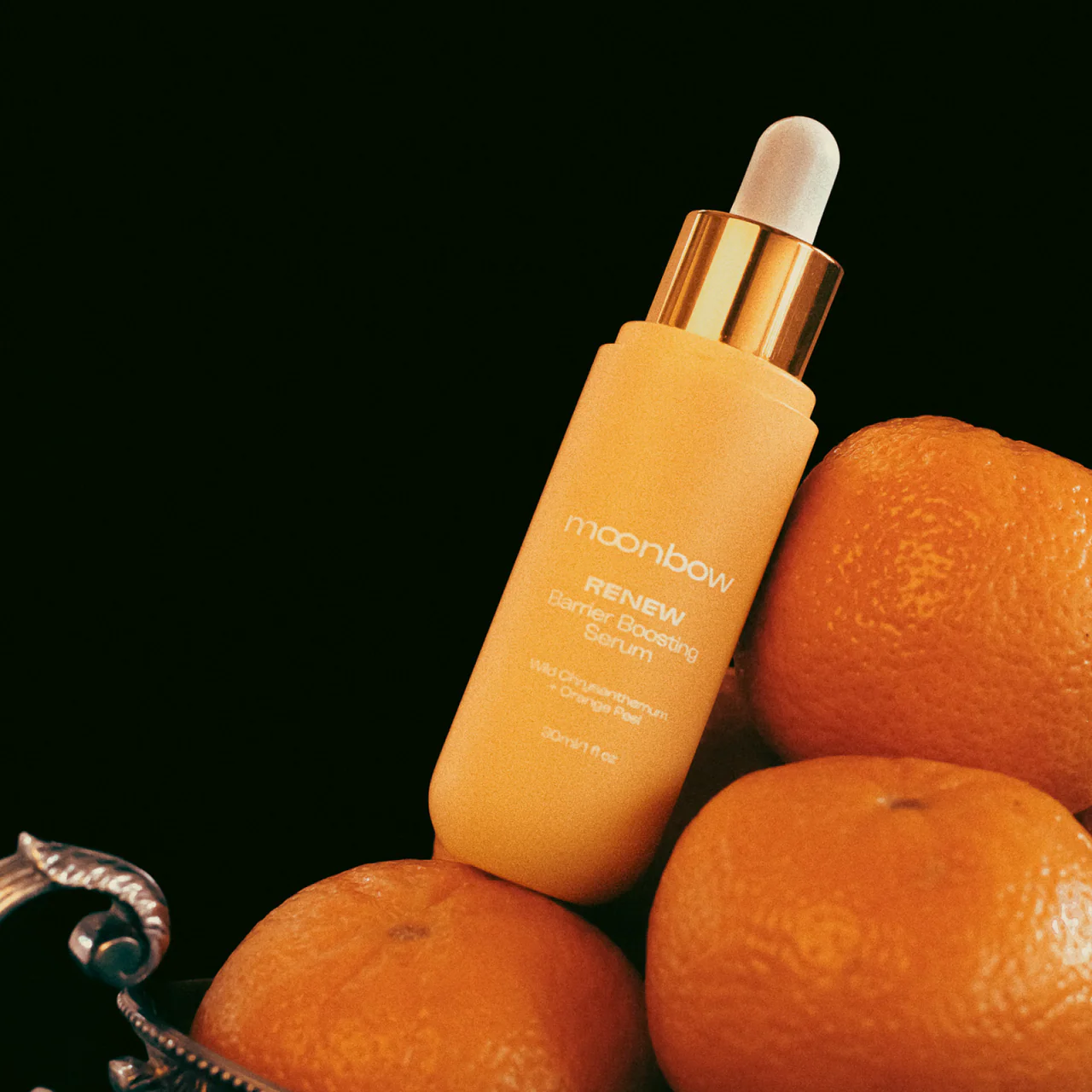 A Beauty Inside & Out Box dropper bottle of Moonbow Renew Barrier Boosting botanical serum, vibrant orange in color, sits among fresh oranges against a dark backdrop, reflecting the harmony and balance inspired by Traditional Chinese Medicine.