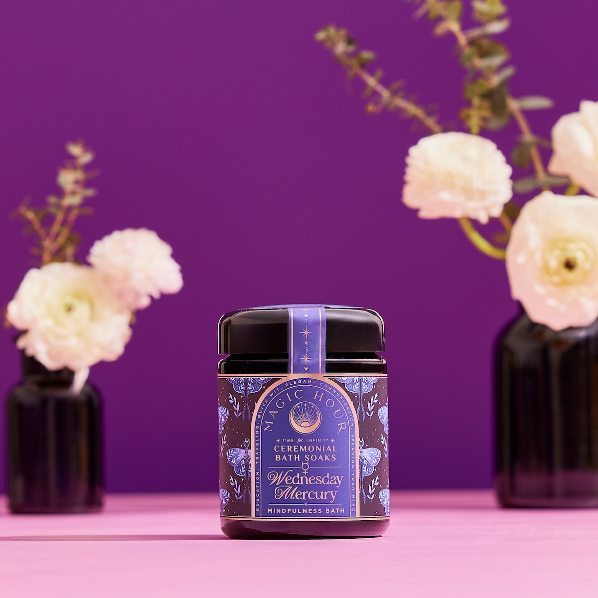A dark violet glass jar of Ceremonial Bath Salt Soaks rests on a pink surface, exuding celestial healing energy, with white flowers in black vases against a vibrant purple backdrop.