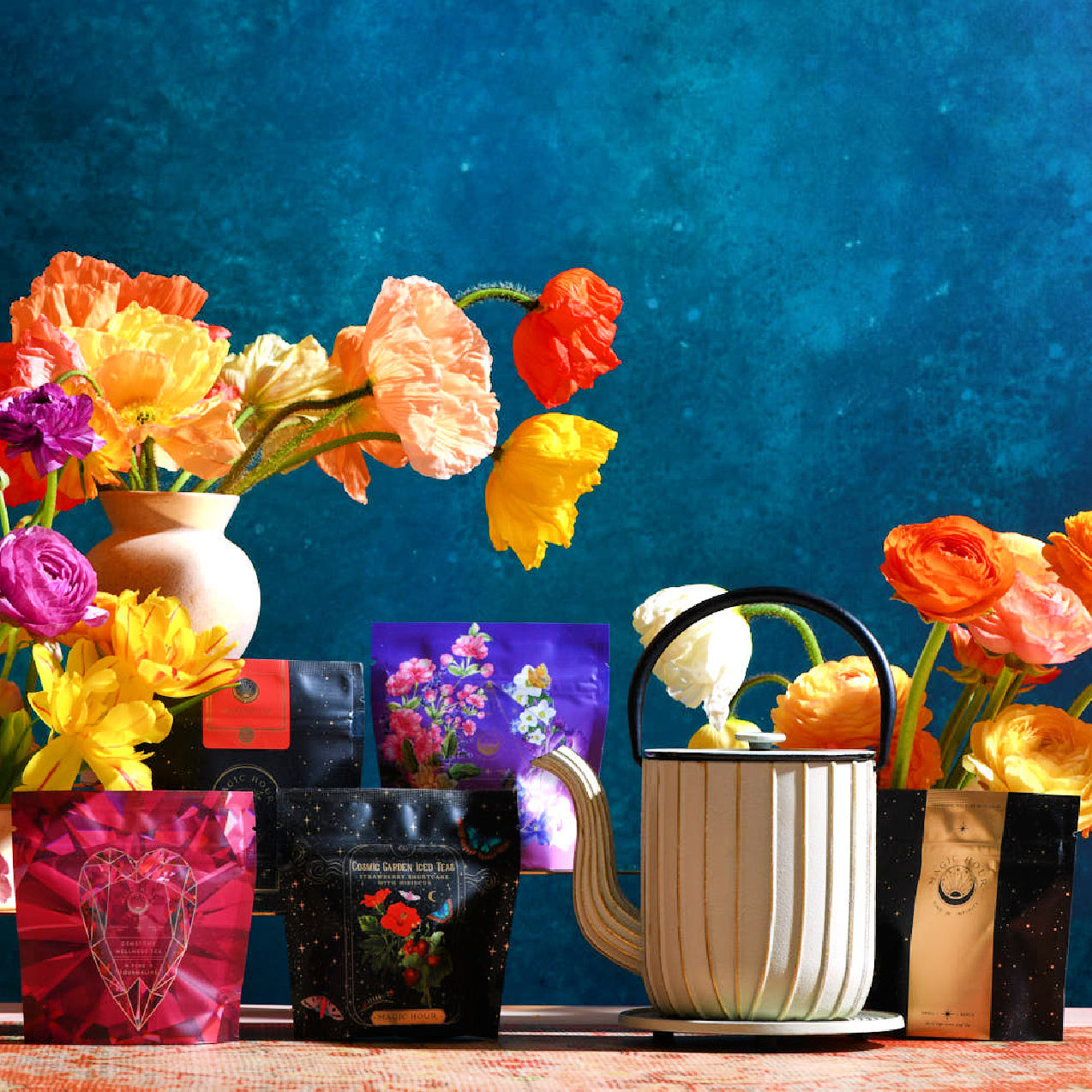 A brightly colored arrangement of flowers in vases is displayed against a vivid blue background. Various colorful packets, possibly seeds or the White Magic Sampler Box from Club Magic Hour, are positioned around a cream-colored, ribbed watering can. The scene exudes a lively and vibrant atmosphere.