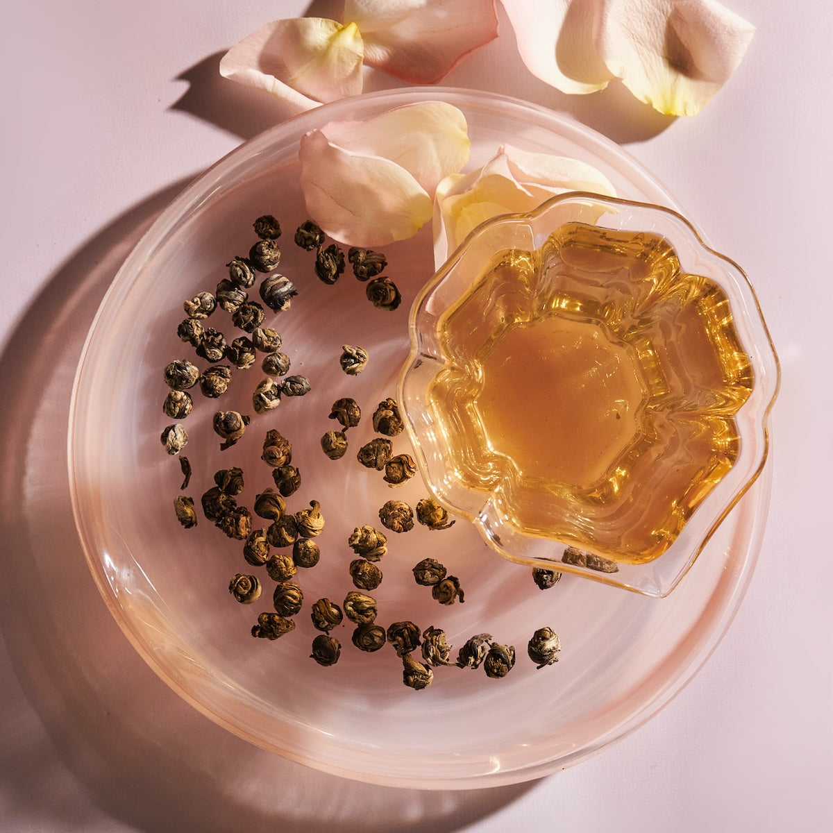 A glass cup of tea sits on a light pink plate, with hand-rolled white tea leaves scattered nearby. Pink flower petals are placed around the plate, creating a delicate arrangement. The lighting casts soft shadows, adding a warm and inviting ambiance that complements the exquisite taste of Magic Hour White Tea Pearls.