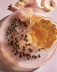 A glass cup of tea sits on a light pink plate, with hand-rolled white tea leaves scattered nearby. Pink flower petals are placed around the plate, creating a delicate arrangement. The lighting casts soft shadows, adding a warm and inviting ambiance that complements the exquisite taste of Magic Hour White Tea Pearls.