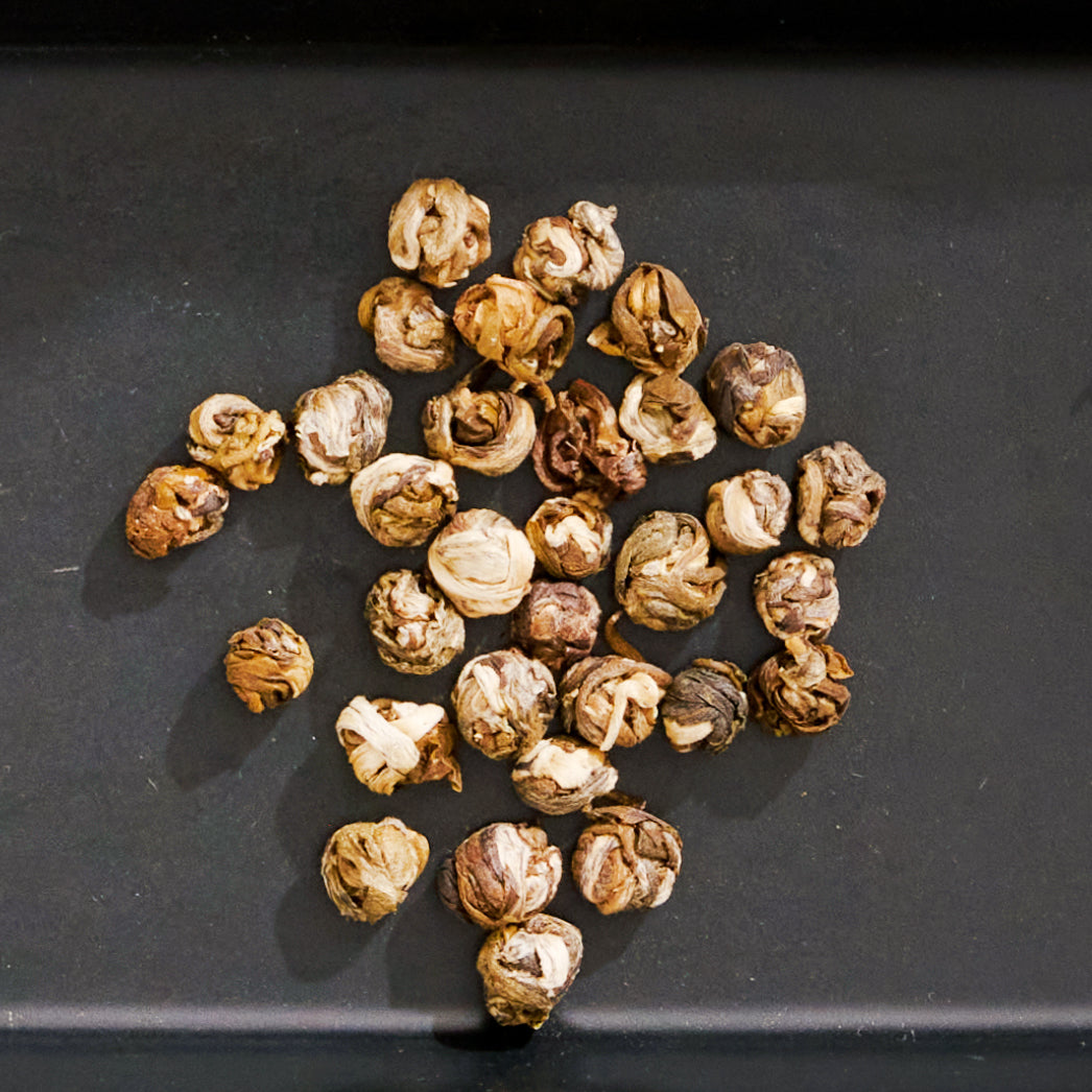 A cluster of dried jasmine flowers is displayed on a dark flat surface. The naturally curled round shapes, reminiscent of White Tea Pearls from Magic Hour, show variations of brown and beige hues, hinting at their exquisite taste.
