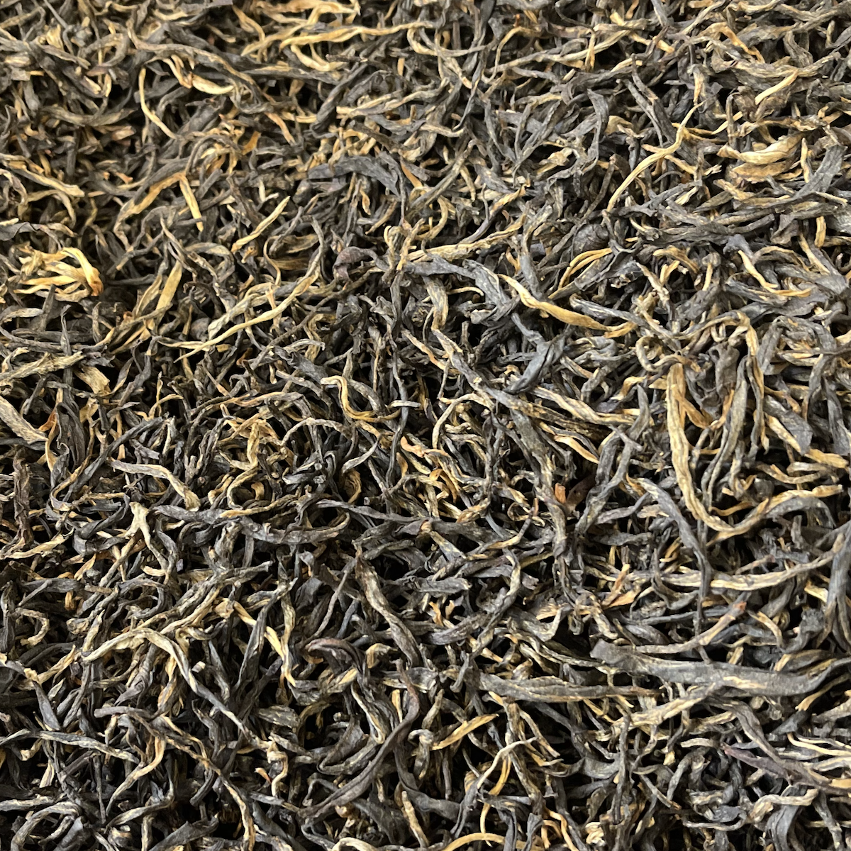 A close-up of Yunnan Ancient Tree Black Tea shows loose, dried leaves with dense dark brown and golden yellow strands, sourced from ancient tea trees.