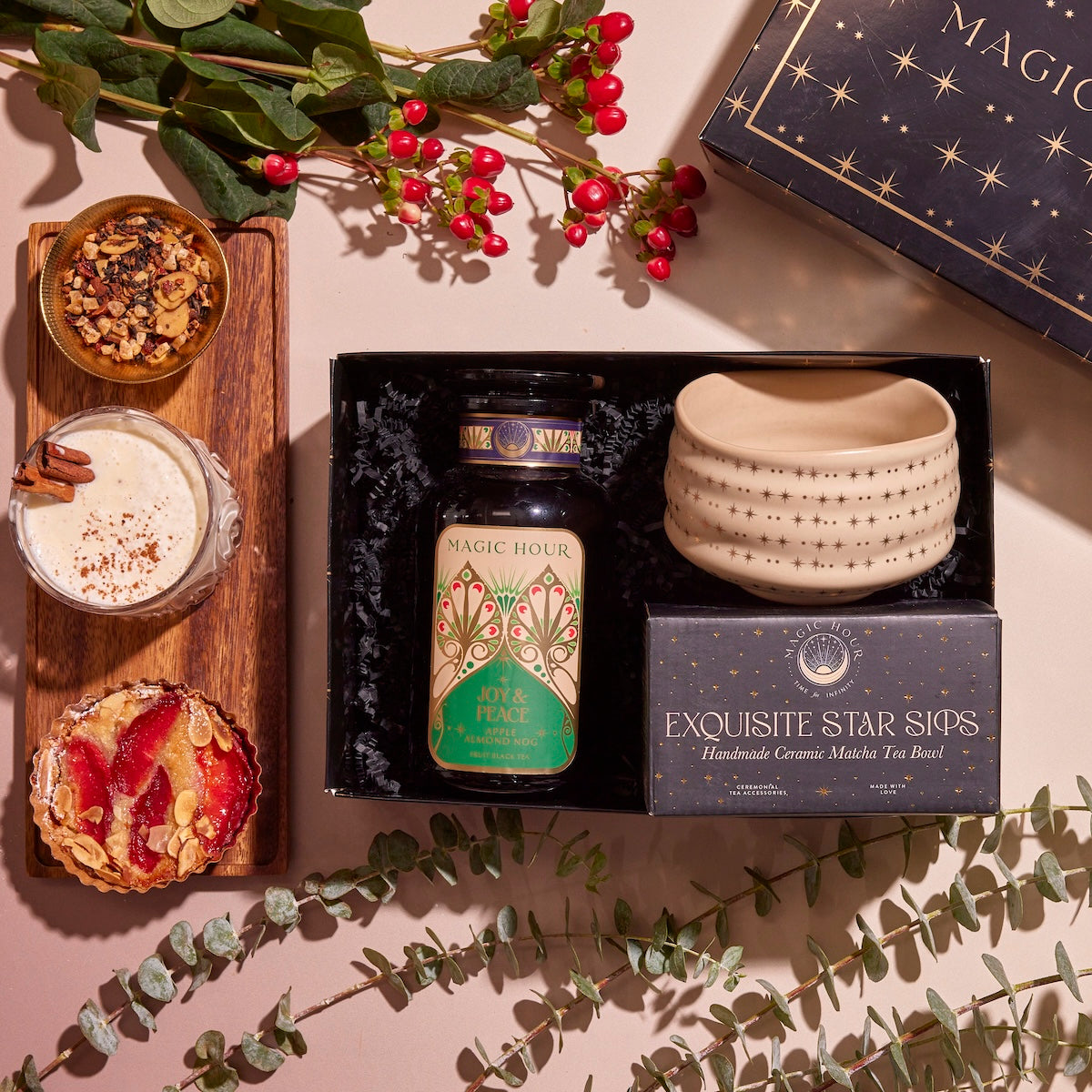 The Winter Wellness Wishes Tea & Accessory Gift Set includes a ceramic mug, herbal tea, and a box labeled "Exquisite Star Sips." Nearby, you'll find a wooden board featuring a creamy drink garnished with cinnamon and two small tarts. Eucalyptus leaves and red berries are artfully scattered around to enhance the presentation.