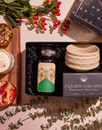 The Winter Wellness Wishes Tea & Accessory Gift Set includes a ceramic mug, herbal tea, and a box labeled "Exquisite Star Sips." Nearby, you'll find a wooden board featuring a creamy drink garnished with cinnamon and two small tarts. Eucalyptus leaves and red berries are artfully scattered around to enhance the presentation.