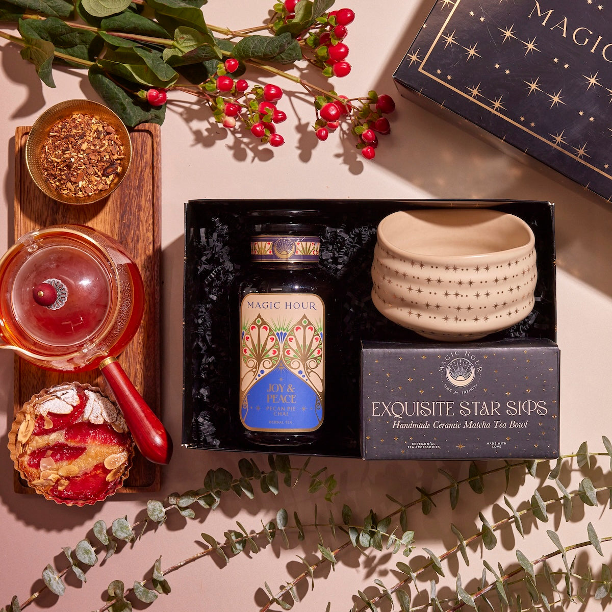 A flat lay of the "Winter Wellness Wishes Tea & Accessory Gift Set" features a luxury gift box containing a jar of seasonal teas, a ceramic tea bowl, and a pot on a wooden tray. It's beautifully complemented by red and yellow bread and decorative greenery with red berries.