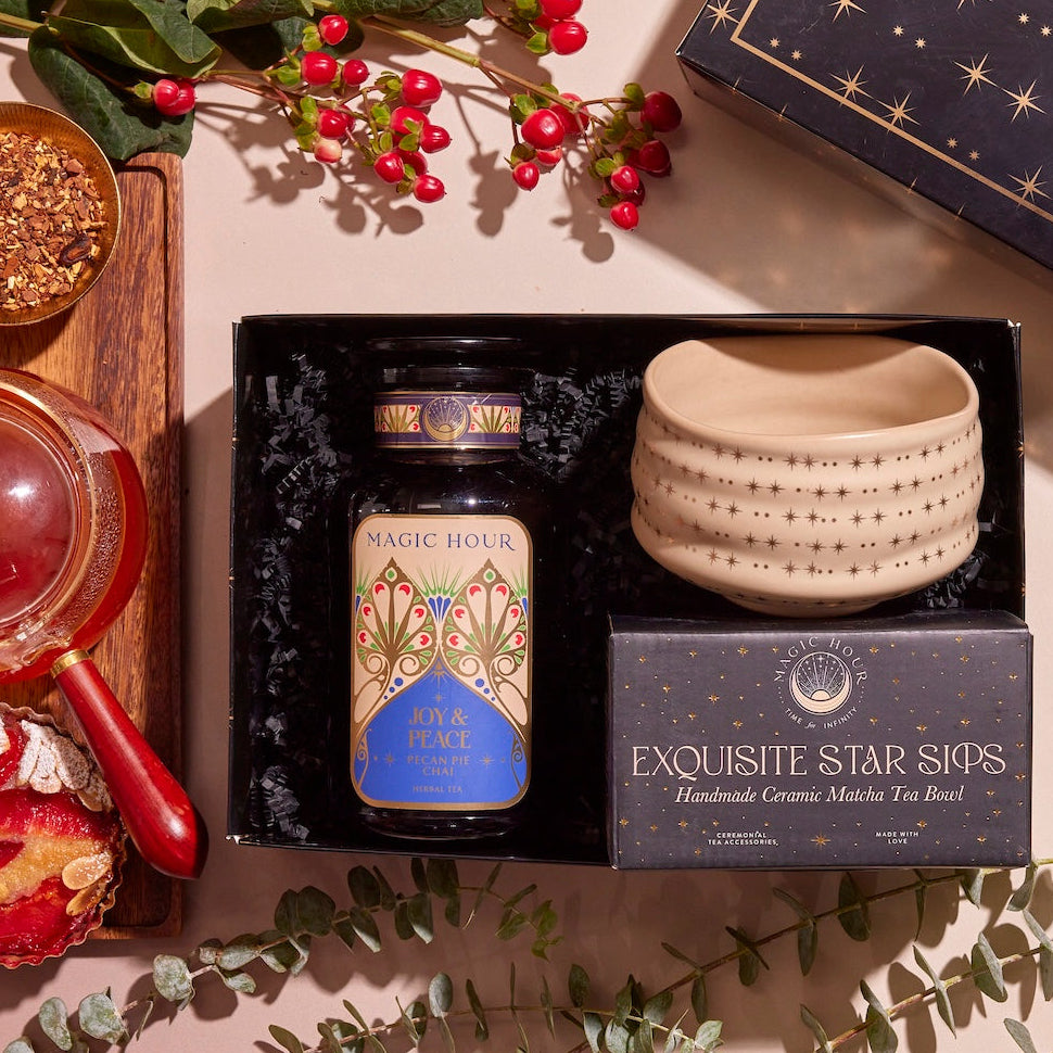 A flat lay of the "Winter Wellness Wishes Tea & Accessory Gift Set" features a luxury gift box containing a jar of seasonal teas, a ceramic tea bowl, and a pot on a wooden tray. It's beautifully complemented by red and yellow bread and decorative greenery with red berries.