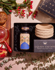 A flat lay of the "Winter Wellness Wishes Tea & Accessory Gift Set" features a luxury gift box containing a jar of seasonal teas, a ceramic tea bowl, and a pot on a wooden tray. It's beautifully complemented by red and yellow bread and decorative greenery with red berries.