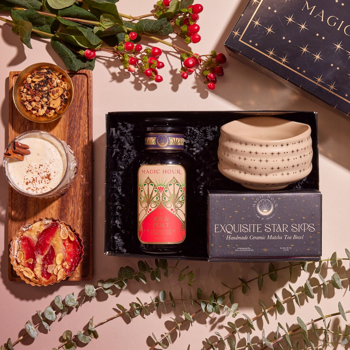 A flat lay of the Winter Wellness Wishes Tea &amp; Accessory Gift Set showcasing a decorated tea jar, a handmade ceramic matcha tea bowl, and a box. The arrangement is surrounded by a wooden board topped with granola, seasonal teas, a latte, a tart, eucalyptus leaves, and red berries.