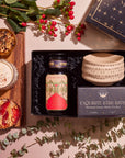 A flat lay of the Winter Wellness Wishes Tea & Accessory Gift Set showcasing a decorated tea jar, a handmade ceramic matcha tea bowl, and a box. The arrangement is surrounded by a wooden board topped with granola, seasonal teas, a latte, a tart, eucalyptus leaves, and red berries.