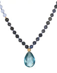 A Magic Hour Wise Spirit Sky Blue Topaz Gemstone Drop Mala with alternating dark-colored and light blue beads, featuring a large, teardrop-shaped, faceted blue topaz gemstone pendant at the center, connected by two small golden beads.