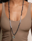 A person wearing a beige sleeveless top displays a Wise Spirit Sky Blue Topaz Gemstone Drop Mala by Magic Hour with alternating black and white beads. The necklace, ideal for meditation practices, features a large, transparent teardrop-shaped pendant. The serene background is a plain light-colored wall.