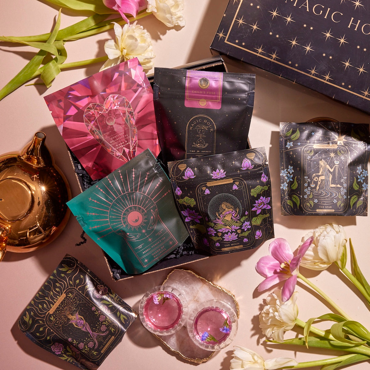 An assortment of elegantly designed tea packages promoting balance is displayed on a surface, surrounded by fresh flowers, a gold teapot, and a black box labeled "Magic Hour." Two pink tea cups and a small bowl with a white rim are also included in the Women's Wellness Sampler Set arrangement.