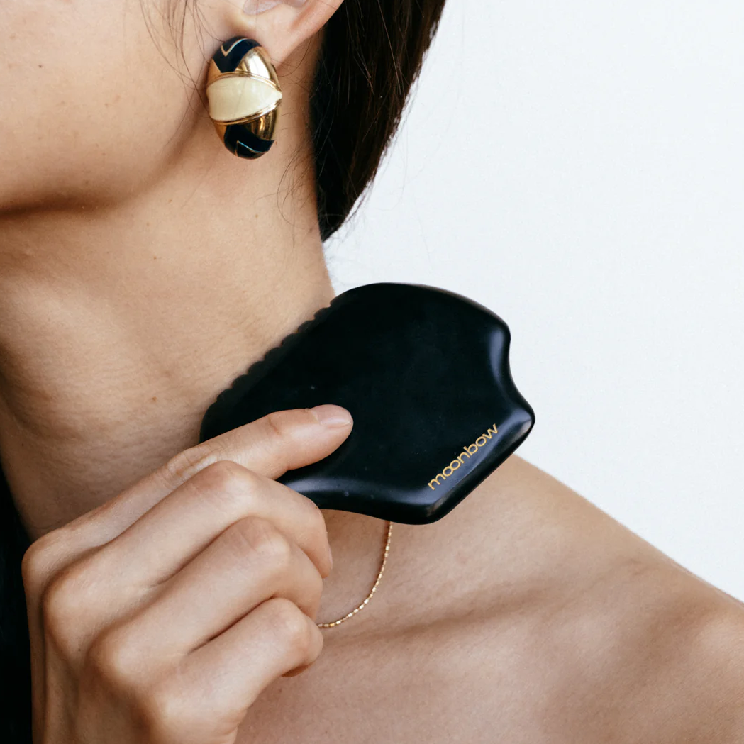 A woman with dark hair uses a black gua sha tool from the Beauty Inside & Out Box, inspired by Traditional Chinese Medicine, holding it to her neck while wearing gold and black earrings and a thin gold necklace.