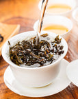 Hot water is being poured over Zhangping Shuixian Oolong loose leaf tea in a white cup with a saucer, with more tea cups and a plate of loose Zhangping Shuixian Oolong leaves in the background.