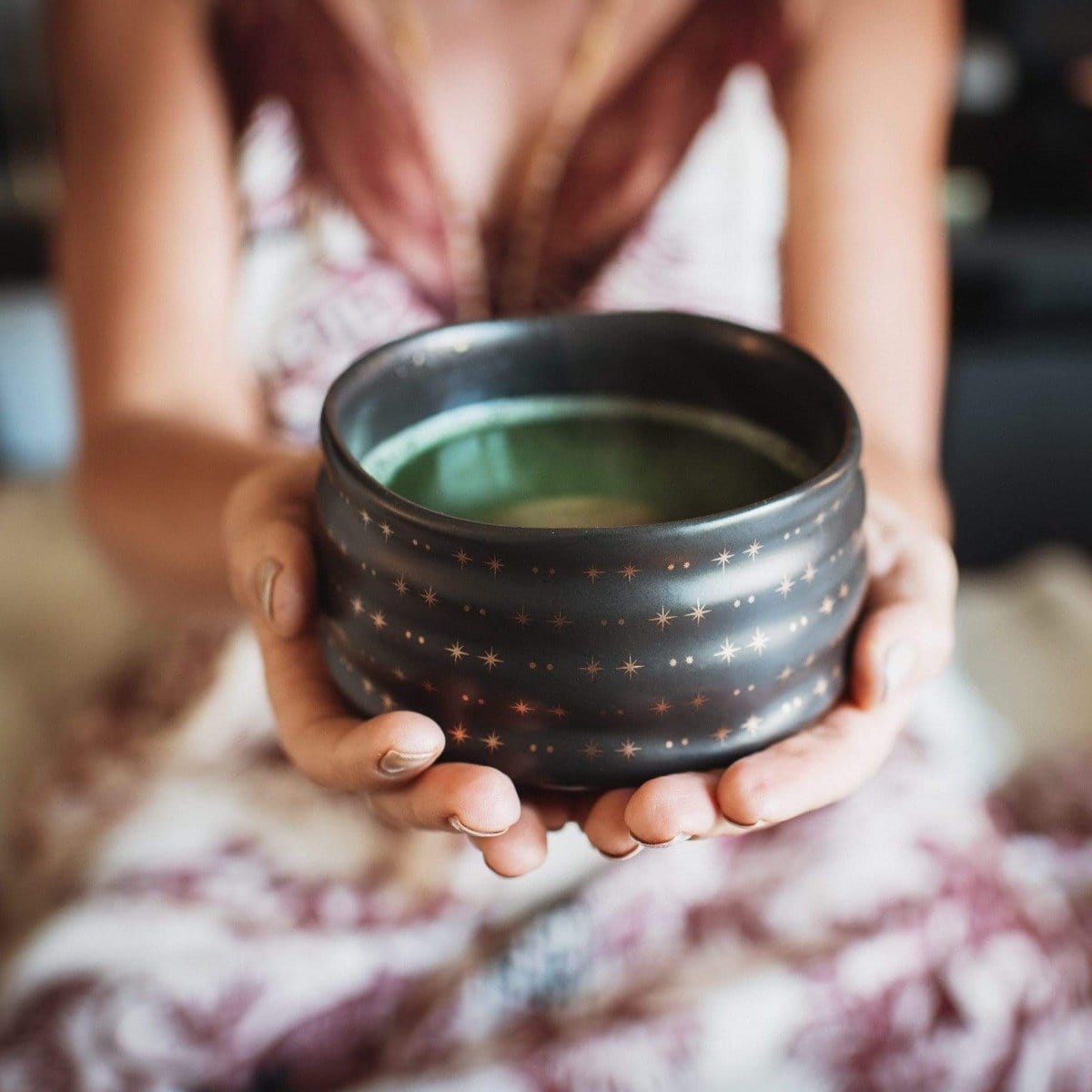 A person holding a black, star-patterned ceramic bowl filled with green liquid from the Magic Hour Starter Kit by Magic Hour. The person is dressed in a light-colored, patterned top or dress and is seated with the bowl held out in both hands. The background is blurred and out of focus.