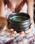 A person holding a black, star-patterned ceramic bowl filled with green liquid from the Magic Hour Starter Kit by Magic Hour. The person is dressed in a light-colored, patterned top or dress and is seated with the bowl held out in both hands. The background is blurred and out of focus.