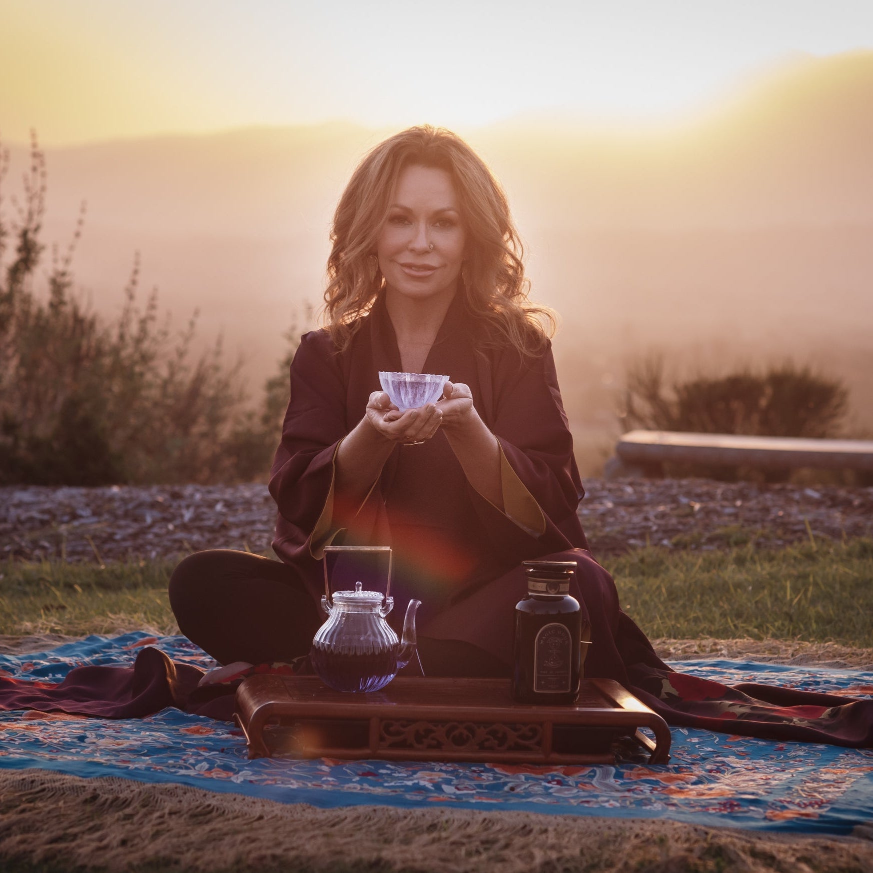 A woman sits cross-legged on a blue and brown patterned mat outdoors at sunset. She holds a teacup with both hands, and in front of her are a glass teapot and a jar on a wooden tray. She has long hair and wears a dark robe. The background is a serene, mountainous landscape.