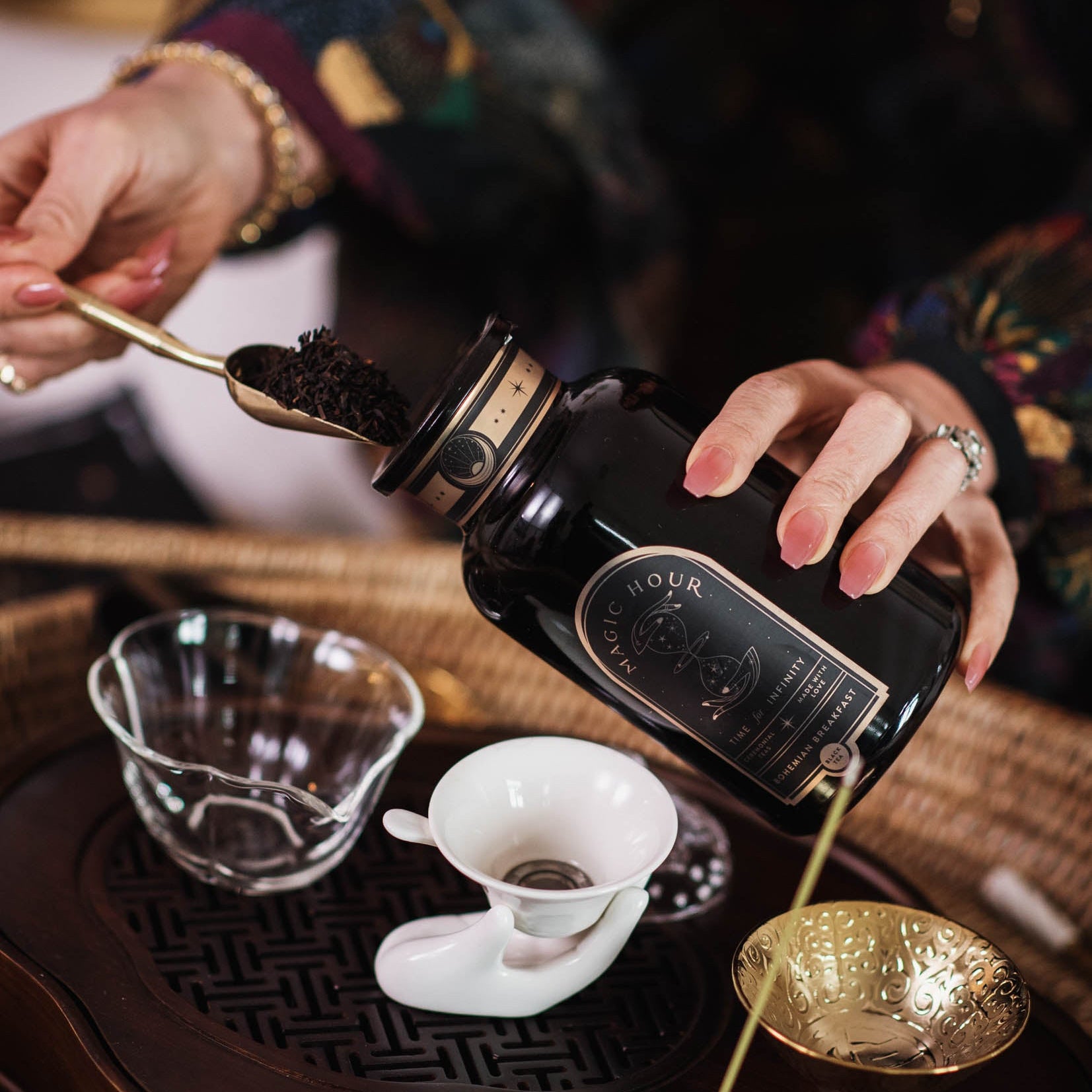 A person scoops loose leaf tea from a black jar into a glass cup on a wooden tray with teaware, featuring Tea Essentials and the In Person & Virtual Tea School Course at the Ojai Tea Salon.