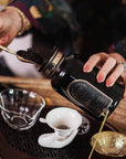 A person scoops loose leaf tea from a black jar into a glass cup on a wooden tray with teaware, featuring Tea Essentials and the In Person & Virtual Tea School Course at the Ojai Tea Salon.