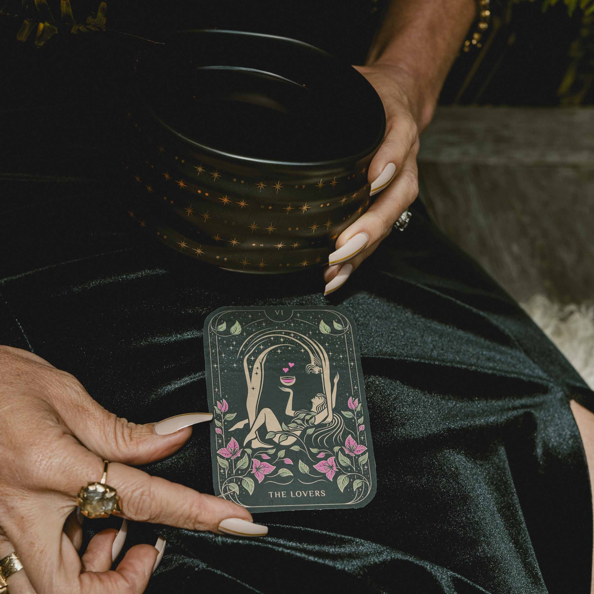 A person with manicured nails holds a black cup adorned with gold stars and displays “The Lovers” card from the Magic Hour 12 Archetype Set on their lap, dressed in a black satin outfit and wearing statement rings.
