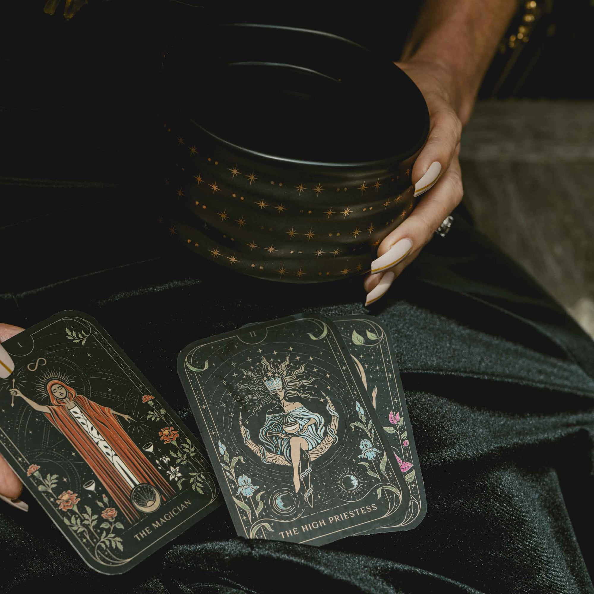 A person holds a black bowl with gold stars, while two illustrated tarot cards, The Magician and The High Priestess from the Magic Hour 12 Archetype Set, rest on their black-clad lap.