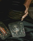 A person holds a black bowl with gold stars, while two illustrated tarot cards, The Magician and The High Priestess from the Magic Hour 12 Archetype Set, rest on their black-clad lap.