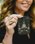 A woman with wavy brown hair, wearing a black outfit and several rings, holds up a tarot card labeled "The Oracle" with an intricate design featuring a meditative figure, celestial elements, and floral motifs. Her face is partially visible and she is smiling; the image evokes tranquility much like the Monthly Magic First Sips Tea Subscription Box from Magic Hour.