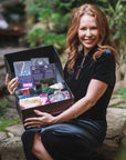 A woman with long, wavy red hair sits outdoors on a rock, smiling and holding an open gift box filled with various colorful items and small snack packages. She is wearing a black dress, and the background is lush with greenery, reminiscent of Magic Hour's Master Tea Blender's serene garden. Among the treasures in her box is "Tea School Course 