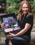 A woman with long red hair, dressed in a dark gown, sits outdoors on a stone step, holding an open black box brimming with a variety of colorful items. She smiles at the camera, surrounded by lush greenery that evokes the serene atmosphere of the Magic Hour Japanese Tea School led by Master Tea Blender Zhena Muzyka, featuring their highly anticipated Tea School 2024-2025 Full Course Bundle.