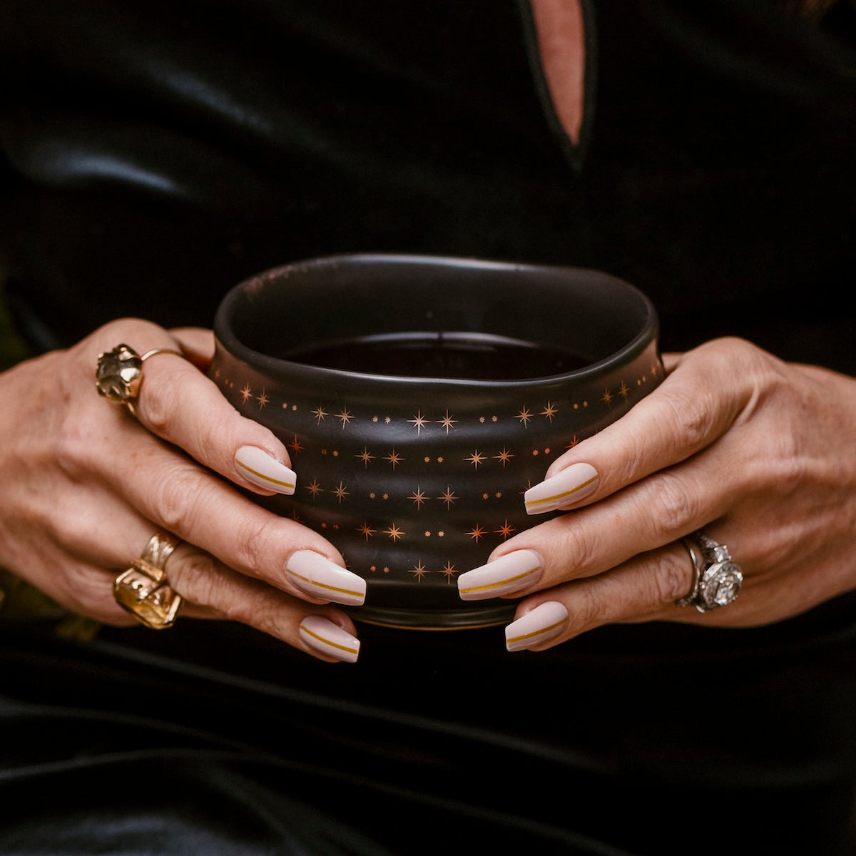 A person with long, manicured nails and multiple rings holds a black School of Magic Monthly Tea Subscription Box - Traveler Jar, adorned with small gold stars, against a dark background.