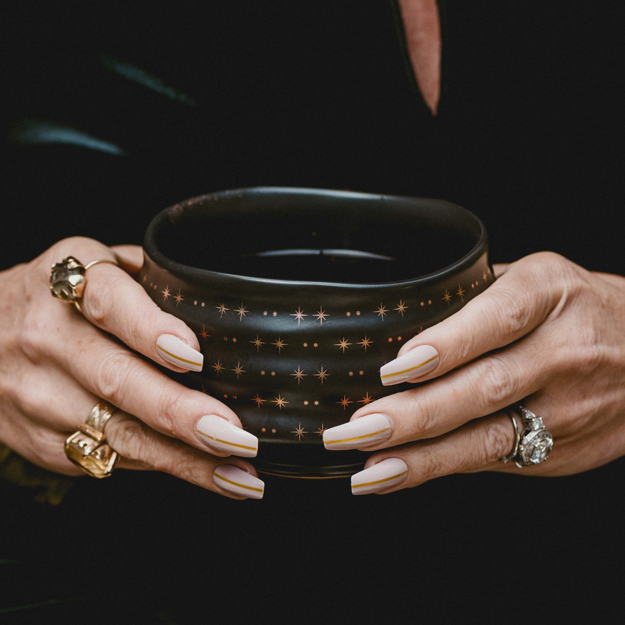 A person with manicured nails and multiple rings holds the Ceremonial Matcha Bowl, a dark cup adorned with delicate star patterns. The mostly black background accentuates the hands and the bowl.