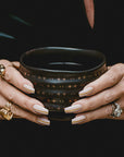 A person with manicured nails and multiple rings holds the Ceremonial Matcha Bowl, a dark cup adorned with delicate star patterns. The mostly black background accentuates the hands and the bowl.