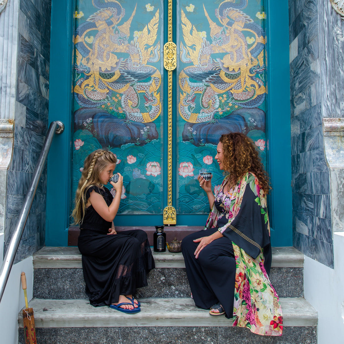 A woman and a girl sit on stone steps in front of an elaborately decorated door featuring dragon artwork. They are both drinking from ceramic cups, with the woman dressed in a colorful, flowing outfit and the girl in a black dress with blue sandals.