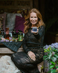 A woman with long, wavy hair, dressed in a black lace outfit, sits on a wooden bench. She smiles and extends a glass of floral tea towards the viewer. The background has a cozy setup with decorative pillows, jars, flowers, and greenery, creating a luxurious and inviting atmosphere—perfect for showcasing Magic Hour's Tea School Course 