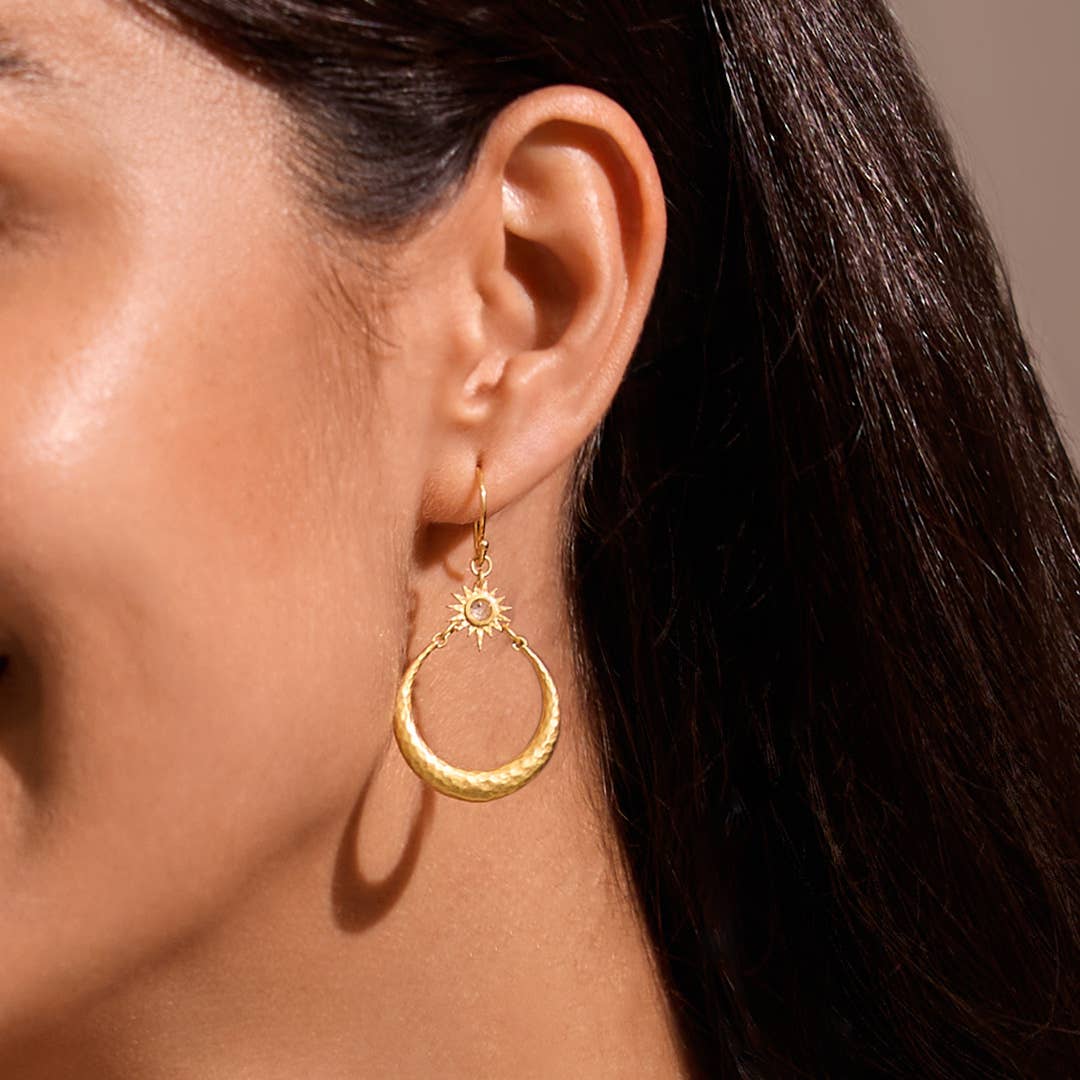 A close-up of a woman's ear wearing Stellar Sisterhood Moonstone Hoops: gold crescent moon earrings with a sun detail and textured finish that catch the light, creating an elegant, celestial-inspired look.