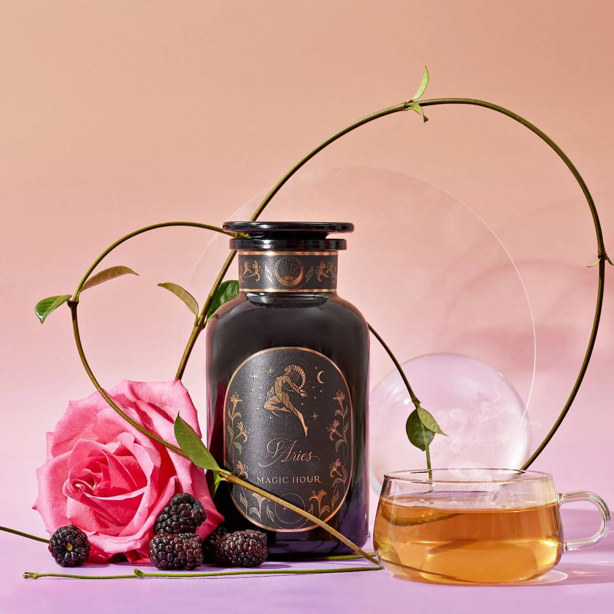 An Aries Astrology Tea Gift Set featuring an apothecary jar labeled “Aries” next to a glass cup of tea, a pink rose, and blackberries.
