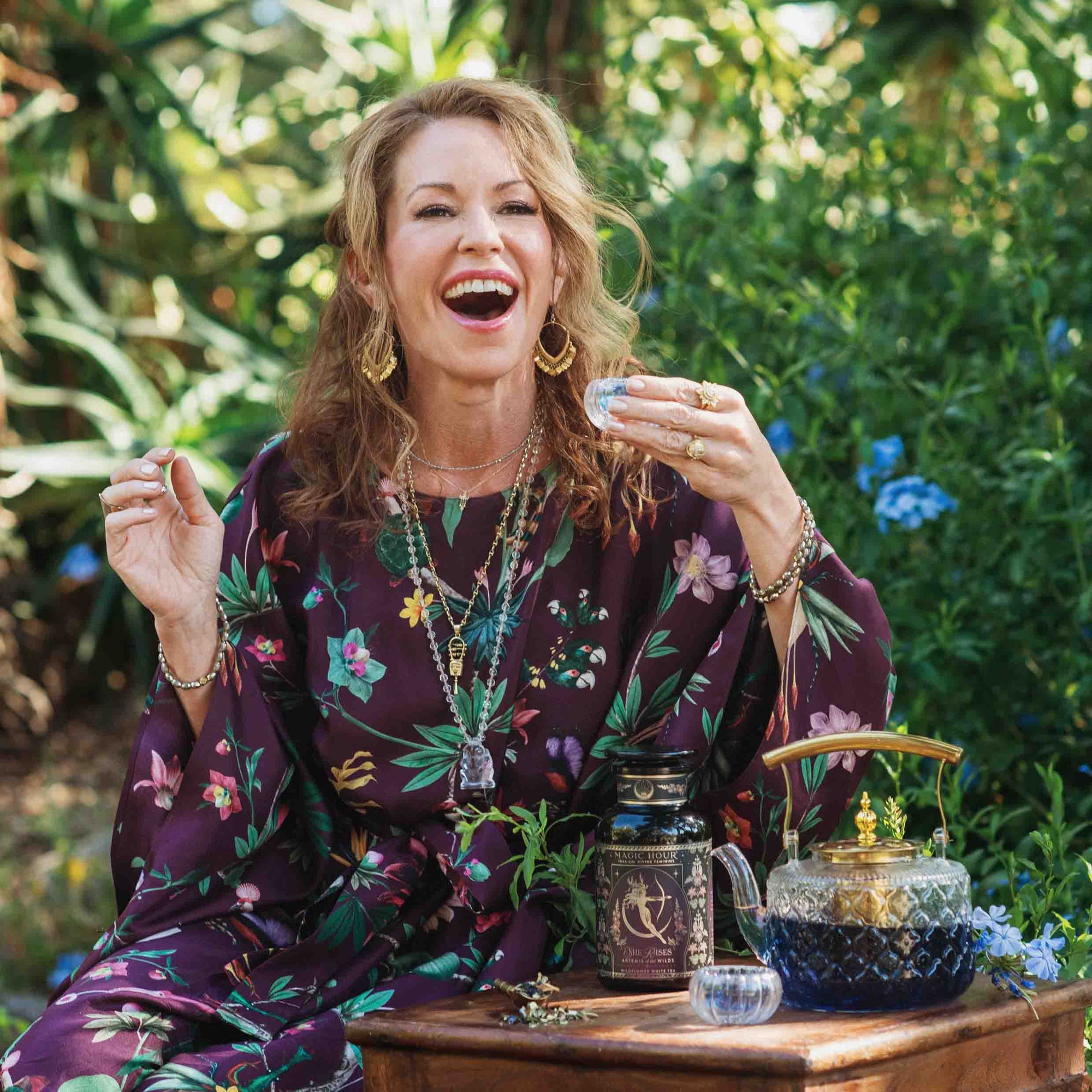 A smiling woman in a floral dress enjoys her Monthly Magic Tea Journey Subscription outdoors, surrounded by greenery and flowers, with a teapot and decorative glass bottles on the wooden table in front of her.