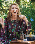 A smiling woman in a floral dress enjoys her Monthly Magic Tea Journey Subscription outdoors, surrounded by greenery and flowers, with a teapot and decorative glass bottles on the wooden table in front of her.