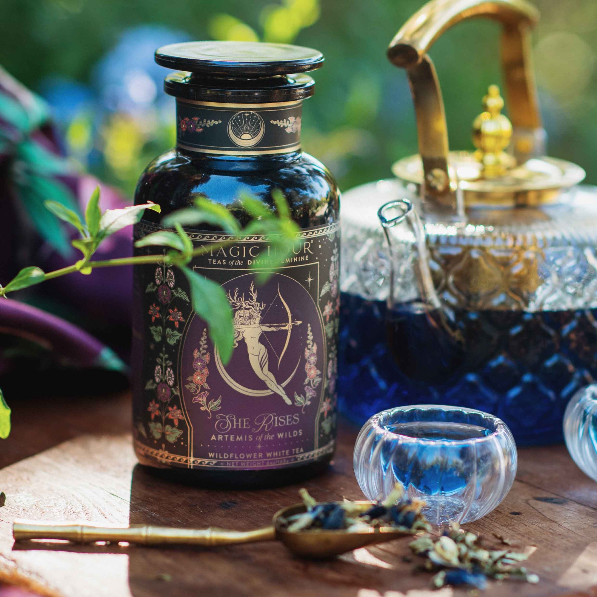 A glass teapot with blue tea, a small glass cup, and a brass teaspoon of loose tea complement the "Monthly Magic Tea Journey Subscription," creating a goddess ritual celebrating the Divine Feminine amid lush greenery.
