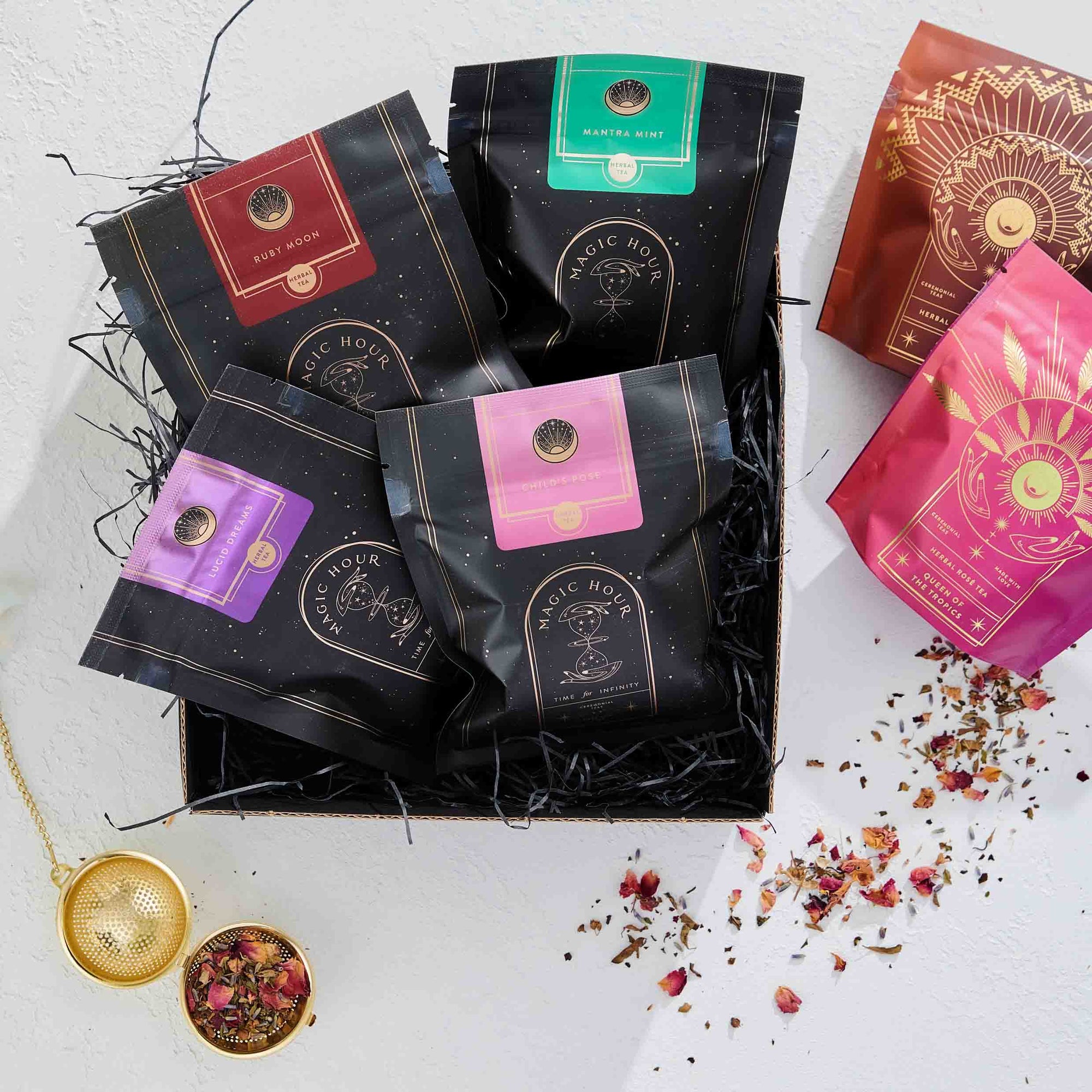 The Herbal Tea Sampler Tea Box offers caffeine-free teas for sleep, digestion & immunity, beautifully arranged with a gold tea strainer and rose petals on black shred, plus two extra tea bags on a white surface.