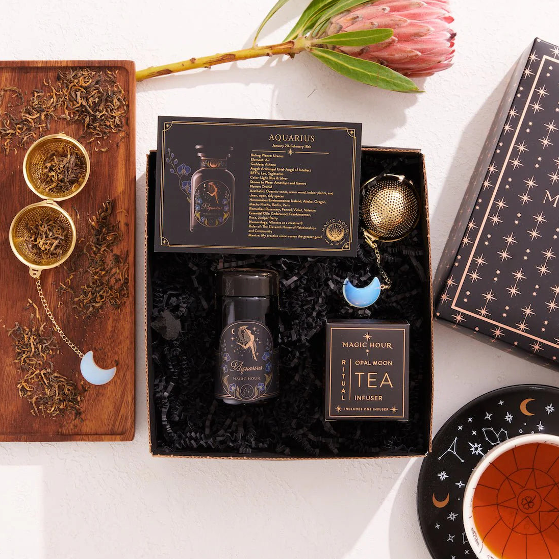 A decorative flat lay showcasing the Aquarius Astrology Tea Gift Set. It features a wooden box filled with loose leaf tea, a black tin labeled "Aquarius," a coaster, a tea infuser adorned with a crescent moon charm, and a protea flower. The starry design elements make it an ideal choice for any Capricorn Gift Set.