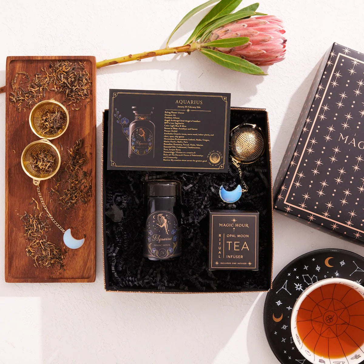 A decorative flat lay showcasing the Aquarius Astrology Tea Gift Set. It features a wooden box filled with loose leaf tea, a black tin labeled &quot;Aquarius,&quot; a coaster, a tea infuser adorned with a crescent moon charm, and a protea flower. The starry design elements make it an ideal choice for any Capricorn Gift Set.