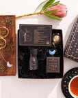 A decorative flat lay showcasing the Aquarius Astrology Tea Gift Set. It features a wooden box filled with loose leaf tea, a black tin labeled "Aquarius," a coaster, a tea infuser adorned with a crescent moon charm, and a protea flower. The starry design elements make it an ideal choice for any Capricorn Gift Set.