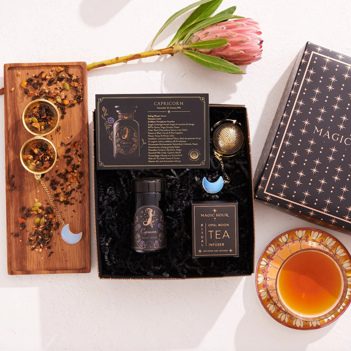 The Capricorn Astrology Tea Gift Set includes a box of tea, a small jar, a tea infuser, and a card titled &quot;Capricorn.&quot; Alongside it is an ornate teacup and an Opalite Moon Tea Strainer placed on a decorative tray embellished with loose tea and a protea flower.