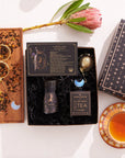 The Capricorn Astrology Tea Gift Set includes a box of tea, a small jar, a tea infuser, and a card titled "Capricorn." Alongside it is an ornate teacup and an Opalite Moon Tea Strainer placed on a decorative tray embellished with loose tea and a protea flower.