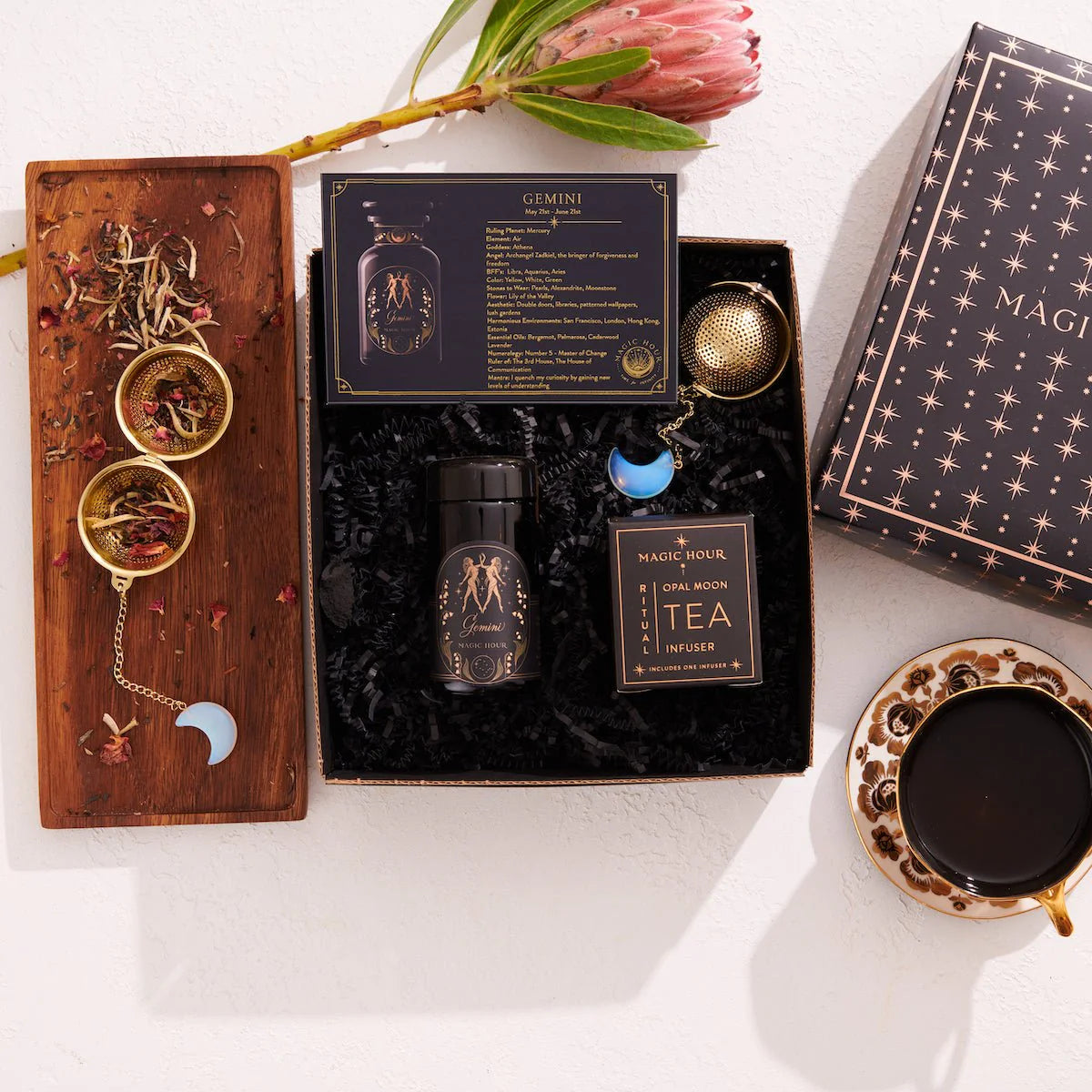 A neatly arranged flat lay showcases a &quot;Gemini Astrology Tea Gift Set,&quot; featuring a tea box, a tea infuser, and a cup of luxury tea on a wooden tray with dried tea leaves and moon-shaped accessories. Soothing botanicals enhance the scene, while a blooming protea flower adds a natural touch against the light background.