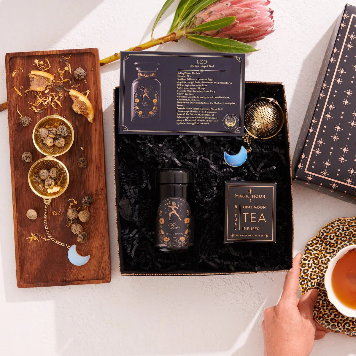 In a flat lay, the Leo Astrology Tea Gift Set is displayed with its vibrant tea blend and an Opalite Moon Tea Strainer. A cup of tea rests next to a wooden tray decorated with chamomile, a crescent moon charm, and dried flowers while a hand reaches for the cup.