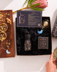 In a flat lay, the Leo Astrology Tea Gift Set is displayed with its vibrant tea blend and an Opalite Moon Tea Strainer. A cup of tea rests next to a wooden tray decorated with chamomile, a crescent moon charm, and dried flowers while a hand reaches for the cup.